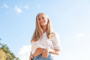 Beautiful young blonde girl with flowing hair and dimples against the blue sky and the sun, pretty smile