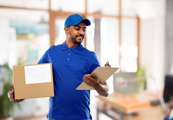 Wall Mural - mail service and shipment concept - happy indian delivery man with parcel box and clipboard in blue uniform over office background