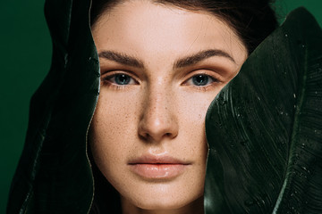 Beautiful girl with freckles on face posing with palm leaves isolated on green