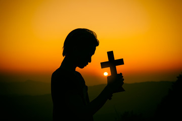 Wall Mural - Young christian praying with holding cross at sunset background. christian silhouette concept.