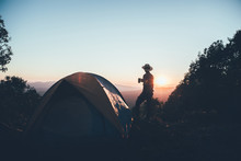Hiker Man In A Hat Standing Holding A Coffee Cup Near Camping Tent On Mountains At Sunset Background. Travel Concept.