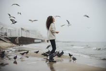 Young Curly Woman Feeding The Birds On The Sea Beach On Foggy Day