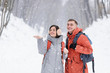 © proimagecontent - Brunette girl catches a snowflake on hand and blonde boy rejoices the snow and smiling to the camera while walking in the snowy forest with backpacks.