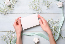 Hands Holding Blank Paper Card On Light Blue Wooden Desk With Flowers. Tender Greeting Card For Womens Or Mothers Day, Easter, Spring Holidays.