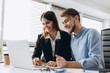 © JJ Studio - Two smiling businesspeople sitting together at a table in a modern office talking and using a laptop