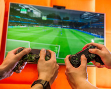 Two Guys Are Enjoying Playing Video Games On The Console. Two Hands And Controller On The Football Or Soccer Background.