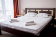 © mirage_studio - Hotel bedroom interior with empty double bed with wooden headboard, bedside table and big window, copy space. White sheet, soft pillows and towels on bed