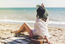 Summer Vacation Concept. Happy Young Woman Relaxing On Beach. Hipster Slim Girl In White Shirt And Hat Sitting And Tanning On Beach Near Sea With Waves, Sunny Warm Weather. Peaceful Calm Moment