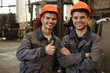 Portrait of two happy workers of metal stock in orange helmets and gray uniform standing together, posing and looking at camera. Man showing sight thumb up and his colleague standing with arms folded.