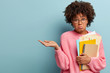 Indoor shot of hesitant African American woman spreads hands, raises palm, feels clueless, holds papers and notepad, dressed in pink sweater, isolated over blue background, copy space aside.