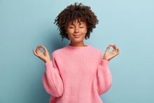 People, Peace And Meditation Concept. Calm Black Young Woman Practices Yoga Indoor, Shows Okay Sign With Both Hands, Demonstrates Approval, Wears Pink Clothing, Isolated Over Blue Background