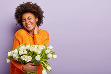 Jubilant Upbeat Black Woman Has Curly Hair, Broad Shining Smile, Keeps Hand On Neck, Eyes Shut, Wears Orange Sweater, Holds White Flowers With Pleasant Scent, Models Over Purple Studio Wall. Spring
