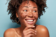 Natural beauty, spa and healthy skin concept. Glad African American woman peels skin with coffee scrub, holds hand on chin, has toothy smile, keeps gaze aside, isolated over blue background.