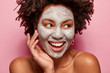 © wayhome.studio  - Photo of glad black woman has clay mask, enjoys facial treatments, keeps gaze aside, touches face, has spa therapy, broad smile with white teeth, isolated over pink background. Perfect skin concept