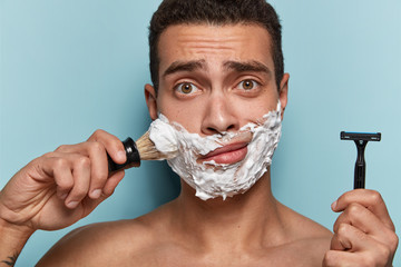 Wall Mural - Men, grooming and shaving concept. Puzzled Caucasian man going to shave beard, holds sharp razor and brush, poses half naked, busy with daily routine in morning, isolated over blue background.