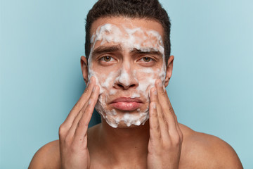 Wall Mural - Image of displeased European man washes face with soap, has discontent expression because of acnes, touches face, poses half nude against blue background in bathroom. Morning hygiene concept