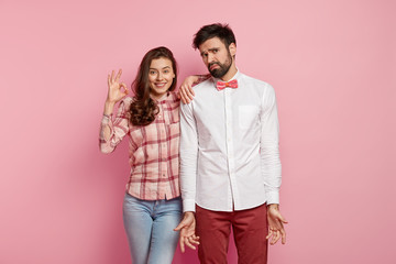 Wall Mural - Horizontal shot of cheerful woman with okay gesture, gives approval, agrees to work together with groupmate who stands clueless, spreads hands, wears elegant clothes, isolated over pink studio wall
