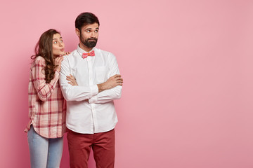 Wall Mural - Thoughtful bearded young man keeps arms folded, wears white shirt with bowtie, being deep in thoughts, beautiful woman in checkered shirt stands closely, keeps lips folded, wants to support.