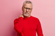Sad elderly man keeps hand near neck, suffers from painful feelings, feels overworked, wears glasses and red jumper, isolated over pink background, wants to sit down or lie. Tiredness, complaining