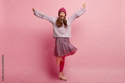 Inspired beautiful overemotive woman clenches fists, spreads hands, stands on one leg, spends time dancing and has fun, wears knitted hat, loose sweater, fashionable skirt, pink pantyhose, celebrates