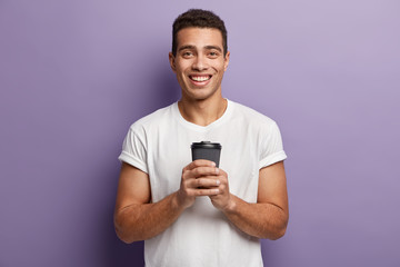 Studio shot of handsome smiling hipster guy with pleased expression, dressed in casual white t shirt, holds takeaway coffee, enjoys hot beverage, isolated over purple background. Drinking concept