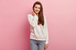 Isolated shot of positive young woman keeps hand on neck, looks directly at camera with pleasant toothy smile, shows white teeth, dressed in casual jumper and jeans, isolated over pink background