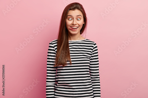 Funny pretty woman sticks out tongue, crosses eyes, plays fool, pretends being silly, wears striped sweater, isolated over pink background. Playful girl feels crazy and amused, makes grimace