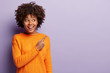 © wayhome.studio  - People, promotion and ethnicity concept. Cheerful dark skinned woman with Afro hairdo, toothy smile, dressed in orange jumper, points with fore finger at upper right corner, poses over lilac wall