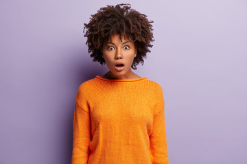 Wall Mural - Stupefied emotional woman with Afro hairstyle, opens mouth with amazement, dressed in orange jumper, looks directly at camera, models over purple background. Human facial expressions concept