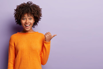 Wall Mural - Glad girl with crisp black hair, dark skin, toothy smile, points with thumb aside, demonstrates something, shows copy space for your usage, wears orange jumper, isolated over purple background