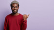 Satisfied black guy with stubble, has toothy broad shining smile and white teeth, wears headgear and red jumper, points with thumb aside, shows blank space for your advertising content or promotion