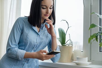 Wall Mural - Morning portrait of young smiling woman in shirt at home near the window with cup of coffee reading text on smartphone