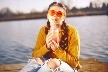 Wall Mural - The girl at hot sunny day drink lemonad. Summer vibes. Girl with fashion yellow sunglasses and dressed in yellow. Beautiful girl sitting on the pier near the river. Beauty model with brunette hair.