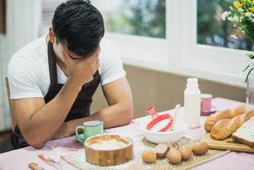 A man sitting which mood strain. On the table have material for prepare dough powder. Chef have a aprons. Live at home in the kitchen.