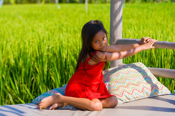 sweet shy and beautiful 7 or 8 years old child in Summer hat and cute red dress having fun outdoors smiling cheerful sitting on resort pool bed at rice field