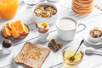 Healthy breakfast eating concept, various morning food - pancakes, soft-boiled egg, toast, oatmeal, granola, fruit, coffee, tea, orange juice, milk on white wooden table