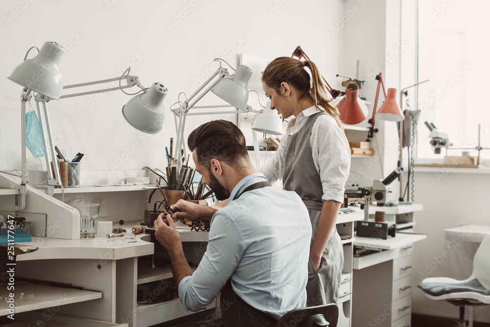 Master and apprentice. Young male assistant and female jeweler are working together at jewelry making workshop. - obrazy, fototapety, plakaty 