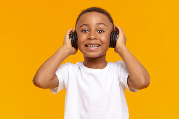 Wall Mural - Isolated shot of funny toothy dark skinned little kid in white t-shirt posing at yellow studio wall in wireless headset, listening to music and smiling, having happy pleased facial expression