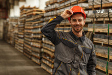 Cheerful Handsome Male Engineer Smiling To The Camera While Putting On Protective Hardhat, Posing At The Storage Of Metalworking Factory. Employment, Industry, Safety Concept