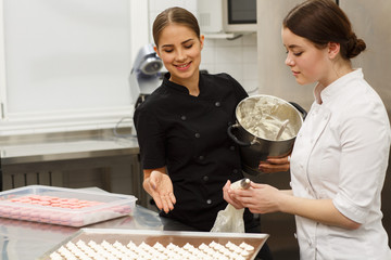 Wall Mural - Female confectioners discussing process of cooking desserts and pointing with hands on baking sheet. Women wearing black and white uniform, one of them holding bowl with sweet cream.