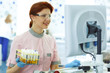 © Nestor - Female lab technician in protective spectacles, pink uniform and blue gloves posing. She holding test tubes with analysis. Woman with ginger hair smiling and looking at computer screen.