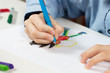Dłonie dziecka trzymają długopis i rysują wymyśloną postać na białej kartce papieru.