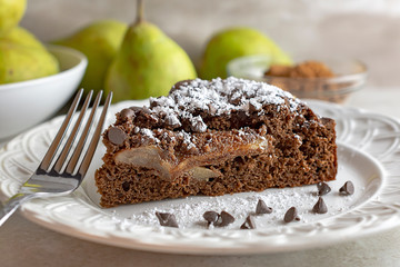 Close up side view of Chocolate Pear Cake sprinkled with powdered sugar.  Fork and chocolate chips on plate.  Pears and cocoa powder in background.