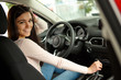 Side view of young smiling female driver sitting in automobile cabin on comfortable seats and posing at camera, turning. Pretty woman holding hand on steering wheel, testing auto.