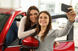 © Nestor - Beautiful and cute girls making selfie near red car. Young female customers smiling and hugging. Girls happy because of buying new automobile in modern car dealership.