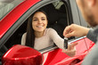 © Nestor - Beautiful and young woman sitting in red car and smiling. Female customer holding her hand and receiving keys of her new car from manager who working in modern car dealership.