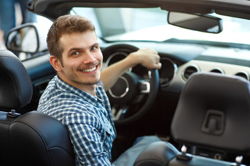 Glad smiling man in checkered shirt and jeans testing new comfortable automobile in car dealership. Handsome customer sitting in car, keeping one hand on wheel, turning back and looking at camera.