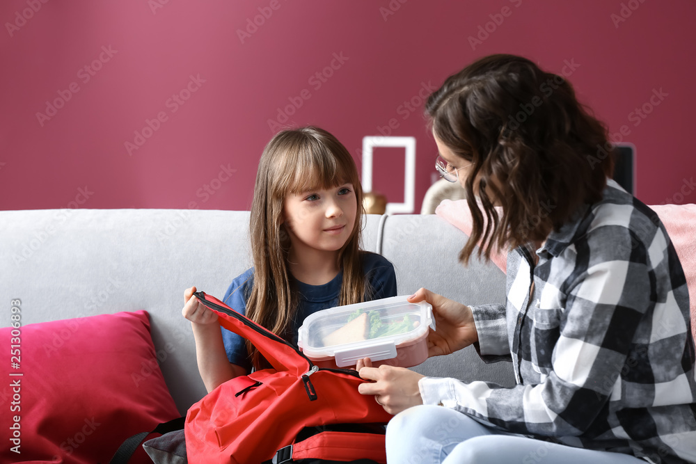 Mother with cute daughter packing lunch box into backpack at home
