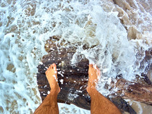 Legs Of A Man Standing On A Snag Lying On The Sandy Seashore Among The Waves And Foam