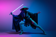 Concept On Cosmic Cosplay. Сontemporary Portrait A Young Athletic Woman In Traditional Japanese Black Kimono, An Asian Hat And Highboots Is Holding A Lightsaber And Posing On Neon Blue-pink Background